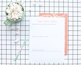 A flat lay composition features a green mug filled with popcorn, placed on a white and black grid-patterned surface. Next to the mug is a worksheet titled 'The Big Idea' with sections for describing a business concept. A mint green pen lies nearby, and some scattered popcorn pieces are visible on the grid.
