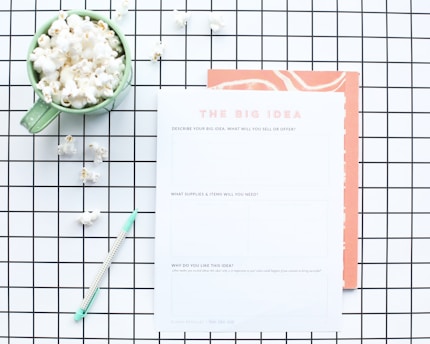 A flat lay composition features a green mug filled with popcorn, placed on a white and black grid-patterned surface. Next to the mug is a worksheet titled 'The Big Idea' with sections for describing a business concept. A mint green pen lies nearby, and some scattered popcorn pieces are visible on the grid.