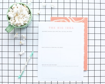 A flat lay composition features a green mug filled with popcorn, placed on a white and black grid-patterned surface. Next to the mug is a worksheet titled 'The Big Idea' with sections for describing a business concept. A mint green pen lies nearby, and some scattered popcorn pieces are visible on the grid.