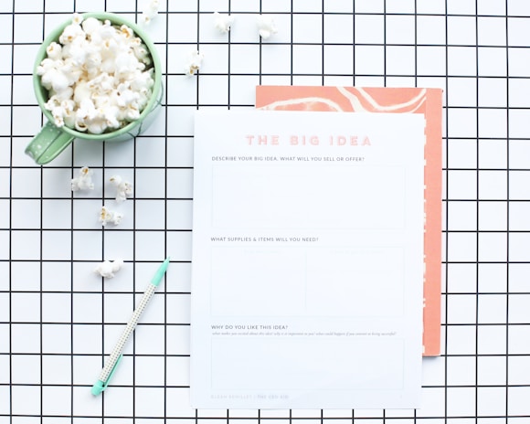 A flat lay composition features a green mug filled with popcorn, placed on a white and black grid-patterned surface. Next to the mug is a worksheet titled 'The Big Idea' with sections for describing a business concept. A mint green pen lies nearby, and some scattered popcorn pieces are visible on the grid.