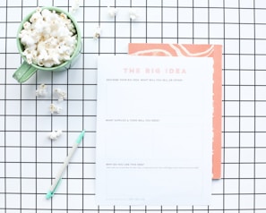A flat lay composition features a green mug filled with popcorn, placed on a white and black grid-patterned surface. Next to the mug is a worksheet titled 'The Big Idea' with sections for describing a business concept. A mint green pen lies nearby, and some scattered popcorn pieces are visible on the grid.