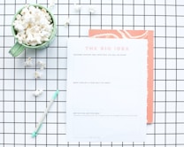 A flat lay composition features a green mug filled with popcorn, placed on a white and black grid-patterned surface. Next to the mug is a worksheet titled 'The Big Idea' with sections for describing a business concept. A mint green pen lies nearby, and some scattered popcorn pieces are visible on the grid.