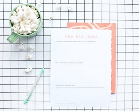 A flat lay composition features a green mug filled with popcorn, placed on a white and black grid-patterned surface. Next to the mug is a worksheet titled 'The Big Idea' with sections for describing a business concept. A mint green pen lies nearby, and some scattered popcorn pieces are visible on the grid.