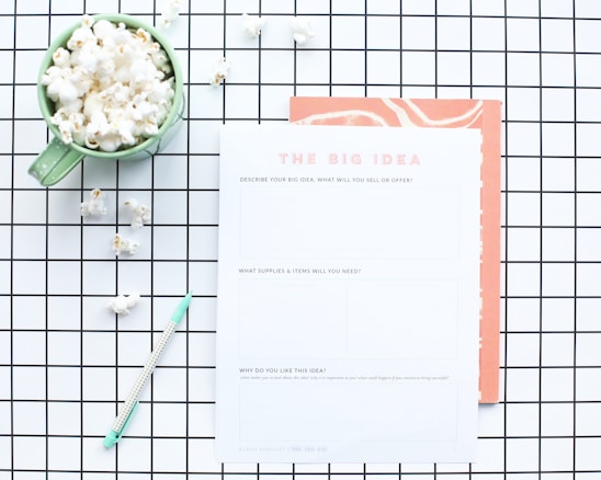 A flat lay composition features a green mug filled with popcorn, placed on a white and black grid-patterned surface. Next to the mug is a worksheet titled 'The Big Idea' with sections for describing a business concept. A mint green pen lies nearby, and some scattered popcorn pieces are visible on the grid.