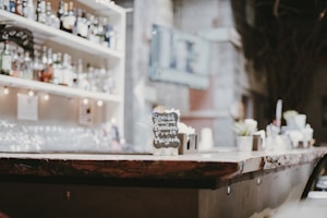 A bar counter with a rustic wooden top, lined with various small plants and a menu sign advertising a special drink. Behind the counter, shelves stocked with a variety of liquor bottles are softly lit, creating a cozy ambiance.