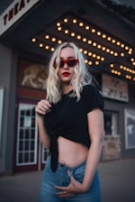 A woman with blonde hair wearing red sunglasses and a black tied-up t-shirt is standing in front of a theater marquee with bright lights.