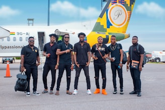 A group of seven men are standing on an airport tarmac in front of a small airplane with a colorful design on its tail. They are casually dressed, most wearing black outfits, and appear to be posing for a group photo. The background includes the airplane with a visible Ghana flag and some airport equipment.