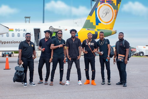 A group of seven men are standing on an airport tarmac in front of a small airplane with a colorful design on its tail. They are casually dressed, most wearing black outfits, and appear to be posing for a group photo. The background includes the airplane with a visible Ghana flag and some airport equipment.