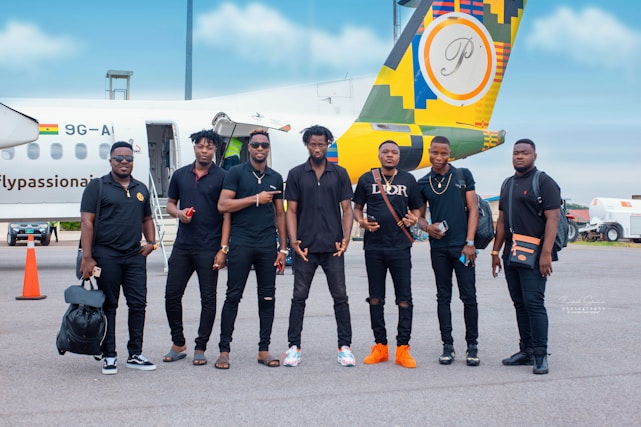 A group of seven men are standing on an airport tarmac in front of a small airplane with a colorful design on its tail. They are casually dressed, most wearing black outfits, and appear to be posing for a group photo. The background includes the airplane with a visible Ghana flag and some airport equipment.