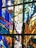 A section of stained glass art featuring a mix of vibrant colors. The design includes decorative elements such as leaves, wheat stalks, and possibly religious motifs. The artwork is arranged in abstract sections with geometric borders.