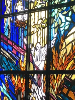 A section of stained glass art featuring a mix of vibrant colors. The design includes decorative elements such as leaves, wheat stalks, and possibly religious motifs. The artwork is arranged in abstract sections with geometric borders.