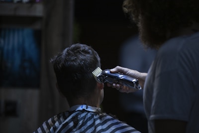 A person with short hair, covered by a barber cape, is having their hair cut with clippers by a barber. The focus is on the clippers and the back of the client's head, while the barber is slightly out of focus. The setting appears to be dimly lit, suggesting a cozy or intimate atmosphere.
