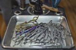 A collection of various scissors and hair cutting tools organized in a metallic tray. The tools are displayed with different colored handles, including gold and iridescent shades. The tray is situated on a dark countertop, with a hairbrush resting nearby.