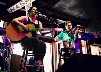 A woman is seated on a stool playing an acoustic guitar and singing into a microphone. Beside her, a man stands playing a violin. Both musicians are illuminated by stage lights, and there is a sign with the word 'NASH' partially visible in the background. The setting appears to be a casual indoor venue with a warm ambiance.