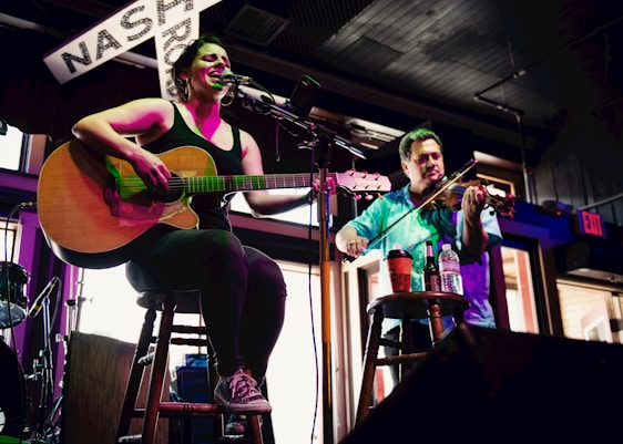 A woman is seated on a stool playing an acoustic guitar and singing into a microphone. Beside her, a man stands playing a violin. Both musicians are illuminated by stage lights, and there is a sign with the word 'NASH' partially visible in the background. The setting appears to be a casual indoor venue with a warm ambiance.