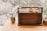 A wooden box with a handle sits on a wooden surface, adorned with a branch bearing round, orange fruits. Beside it, a slender vase holds a single branch with green leaves. The background is gently blurred, showcasing an out-of-focus natural setting.