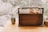 A wooden box with a handle sits on a wooden surface, adorned with a branch bearing round, orange fruits. Beside it, a slender vase holds a single branch with green leaves. The background is gently blurred, showcasing an out-of-focus natural setting.