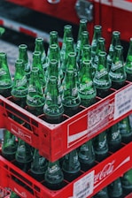 Stacks of green Sprite glass bottles are neatly arranged in red Coca-Cola branded crates. The bottles are empty and arranged vertically, filling each crate. The setting appears to be outdoors, with a blurred background.