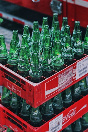 Stacks of green Sprite glass bottles are neatly arranged in red Coca-Cola branded crates. The bottles are empty and arranged vertically, filling each crate. The setting appears to be outdoors, with a blurred background.