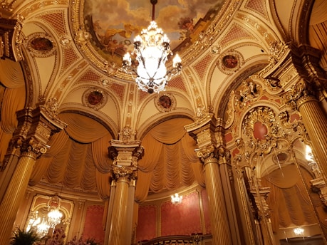 An opulent interior with intricate architectural details, featuring a large chandelier hanging from a decorated, ornate ceiling. The ceiling has elaborate gold moldings and painted murals. Tall, richly adorned columns support the structure, with lavish draperies and gold embellishments throughout.