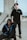 Two individuals dressed in black streetwear are positioned next to a large concrete structure. One is seated on the ground with a blue-accented jacket, while the other stands against the structure, wearing a black T-shirt with a necklace. The background is an outdoor urban environment with a clear sky.