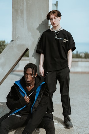 Two individuals dressed in black streetwear are positioned next to a large concrete structure. One is seated on the ground with a blue-accented jacket, while the other stands against the structure, wearing a black T-shirt with a necklace. The background is an outdoor urban environment with a clear sky.