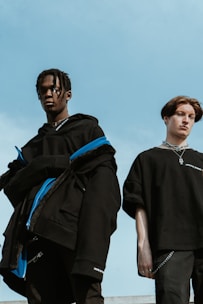 Two young individuals are dressed in modern, dark clothing, standing against a bright blue sky. The person on the left wears a black jacket with blue accents and has their hair styled in short braids. The person on the right is wearing a black shirt and has short, styled hair. Both appear to be posing with a relaxed but serious demeanor.