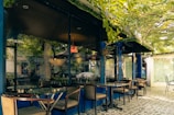 A tranquil outdoor café setting featuring glass tables and wooden chairs under the shade of lush green trees. The reflection in the glass windows shows a calm street scene with plants and a car. The pavement is covered with patterned tiles, adding to the aesthetic appeal.