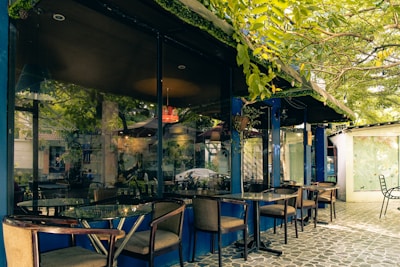 A tranquil outdoor café setting featuring glass tables and wooden chairs under the shade of lush green trees. The reflection in the glass windows shows a calm street scene with plants and a car. The pavement is covered with patterned tiles, adding to the aesthetic appeal.