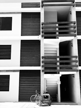 A modern apartment building facade with vertical and horizontal lines. The image is in black and white, highlighting the contrast between the dark shutters and lighter walls. A bicycle with a child trailer is parked in front of the entrance, next to a set of numbers '1794' above the doorway. Small light fixtures are attached to the walls near the entrance.