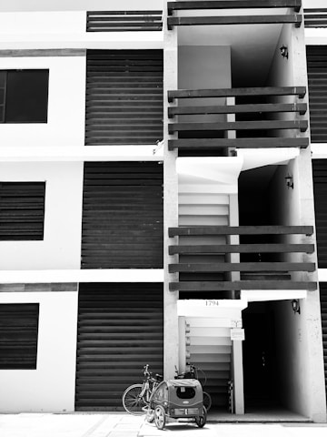 A modern apartment building facade with vertical and horizontal lines. The image is in black and white, highlighting the contrast between the dark shutters and lighter walls. A bicycle with a child trailer is parked in front of the entrance, next to a set of numbers '1794' above the doorway. Small light fixtures are attached to the walls near the entrance.