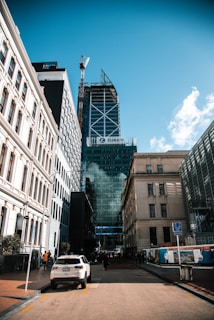 A bustling city street flanked by tall modern and classic buildings. A prominent skyscraper with glass façades features a large sign reading 'Zurich'. A crane is visible at the top of the skyscraper, indicating ongoing construction. A white car is parked on the street, and pedestrians are visible in the distance.