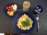 A meal is presented on a table, featuring a blue plate with pasta garnished with greens. Adjacent is another blue plate containing strawberries, apple slices, and a piece of cake. Nearby, there is a tall glass of iced coffee and a shorter glass of iced water. A fork is placed to the right of the plates.