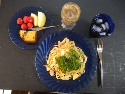 A meal is presented on a table, featuring a blue plate with pasta garnished with greens. Adjacent is another blue plate containing strawberries, apple slices, and a piece of cake. Nearby, there is a tall glass of iced coffee and a shorter glass of iced water. A fork is placed to the right of the plates.