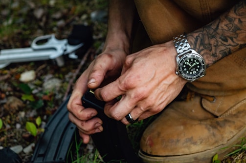 A person's hands loading ammunition into a magazine, with a silver wristwatch and tattoo visible. The person is wearing brown boots, and there are green leaves and a revolver partially visible on the ground.