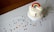 A white cake decorated with a colorful rainbow and clouds made of icing, topped with sprinkles. The cake sits on a white cake stand placed on a table covered with a white cloth. Colorful confetti dots are scattered across the table.