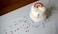 A white cake decorated with a colorful rainbow and clouds made of icing, topped with sprinkles. The cake sits on a white cake stand placed on a table covered with a white cloth. Colorful confetti dots are scattered across the table.
