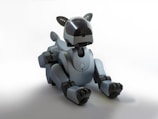 A sleek, futuristic robotic dog with a streamlined design and a matte gray finish sits on the floor. The robot has jointed limbs, a short tail, and a head with a black visor suggesting eyes or sensors. Details like small antennas or ears on the top of the head enhance its animal-like appearance.