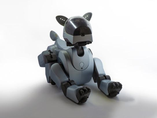 A sleek, futuristic robotic dog with a streamlined design and a matte gray finish sits on the floor. The robot has jointed limbs, a short tail, and a head with a black visor suggesting eyes or sensors. Details like small antennas or ears on the top of the head enhance its animal-like appearance.