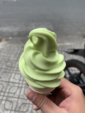 A hand holds a soft serve ice cream cone with a light green swirl against a background of a tiled pavement and a blurred street.