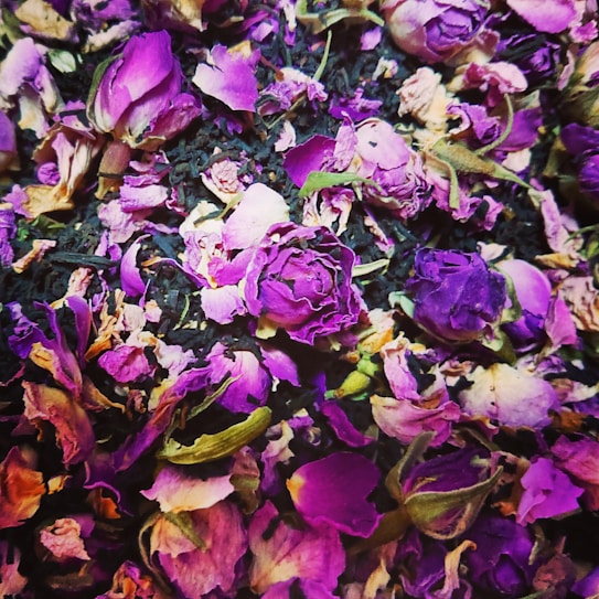 A vibrant collection of dried rose petals and buds in rich shades of purple and pink, layered over a bed of dried tea leaves or herbs. The composition creates a textured, colorful display with a natural, rustic appearance.