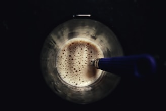 A stainless steel cup containing a frothy beverage, possibly coffee or a milkshake. A blue-handled whisk or stirrer is partially submerged in the drink, with bubbles visible on the liquid's surface. The background is dark, contrasting with the lighter tones of the cup and its contents.