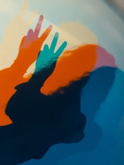 Colorful overlapping shadows of hands making peace signs against a muted background create a playful and artistic composition. The shadows are layered in shades of blue, orange, and pink with a soft, blurred effect, giving it a dynamic yet dreamy appearance.