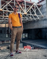 A person stands confidently on a construction site, wearing sunglasses, an orange polo shirt, beige pants, and red sneakers. The background features unfinished concrete structures and metal beams, indicating the area is under construction.