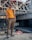 A person stands confidently on a construction site, wearing sunglasses, an orange polo shirt, beige pants, and red sneakers. The background features unfinished concrete structures and metal beams, indicating the area is under construction.