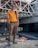A person stands confidently on a construction site, wearing sunglasses, an orange polo shirt, beige pants, and red sneakers. The background features unfinished concrete structures and metal beams, indicating the area is under construction.