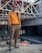 A person stands confidently on a construction site, wearing sunglasses, an orange polo shirt, beige pants, and red sneakers. The background features unfinished concrete structures and metal beams, indicating the area is under construction.