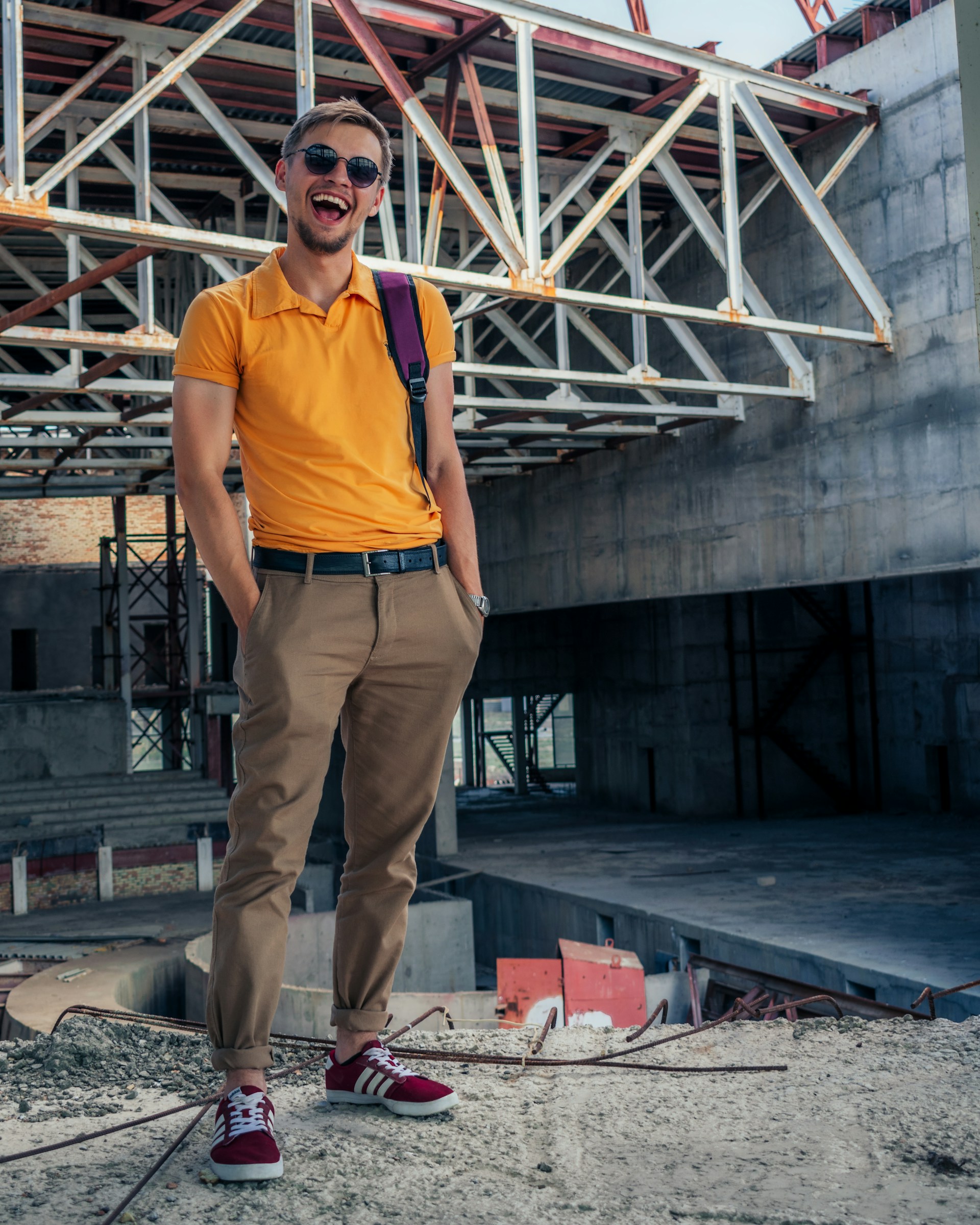 A person stands confidently on a construction site, wearing sunglasses, an orange polo shirt, beige pants, and red sneakers. The background features unfinished concrete structures and metal beams, indicating the area is under construction.