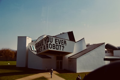 An architectural structure with an abstract design features prominent angular shapes and surfaces. The building's exterior is predominantly white with some black roofing elements. A large sign on the building reads 'HAVE YOU EVER MET A ROBOT?'. A person is seen walking on the path towards the entrance, and there are a few other individuals on the surrounding grass area.