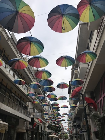 Multicolored umbrellas are suspended in the air above a narrow urban street lined with buildings. The umbrellas create a vibrant canopy that contrasts with the overcast sky. The street is flanked by shops and other establishments, with signs visible on the buildings.